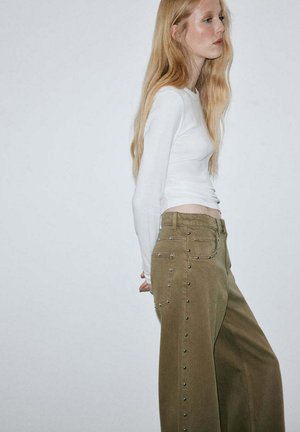 Jeune femme aux longs cheveux blonds portant un haut blanc moulant à manches longues et un pantalon marron ample avec des clous métalliques sur les côtés.