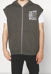 Hombre vestido con sudadera sin mangas de cremallera gris oscuro con texto gráfico en blanco "ACELERACIÓN SIN PARAR" sobre camiseta blanca y pantalones negros.