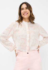 Blusa leggera e trasparente di colore bianco con un delicato motivo floreale rosa, caratterizzata da un colletto alto, maniche lunghe e apertura con bottoni davanti.