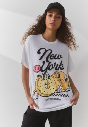 Młoda kobieta w czapce z daszkiem i białej koszulce z kreskówkowymi bajglami z sezamem i napisem "New York", trzymająca brzeg koszulki jedną ręką.