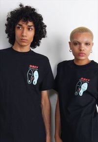 Tricouri negre din bumbac cu un print grafic ce reprezintă tălpi de pantofi, având textul "OBEY" în roșu și accente în alb și verde teal.