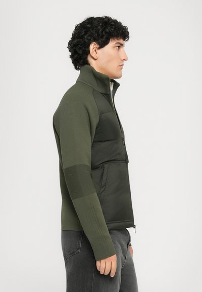 Young man in profile wearing an olive green zip-up jacket with padded front and knit sleeves, paired with dark grey jeans.