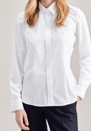 Femme portant une chemise blanche cintrée à manches longues boutonnée et un pantalon noir, debout avec une main dans la poche sur un fond neutre.