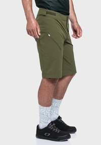 Olivgrüne Shorts aus leichtem Stoff, mit einer seitlichen Reißverschlusstasche und einem Standardbund. Getragen mit gemusterten weißen und grünen Socken und schwarzen Schuhen.
