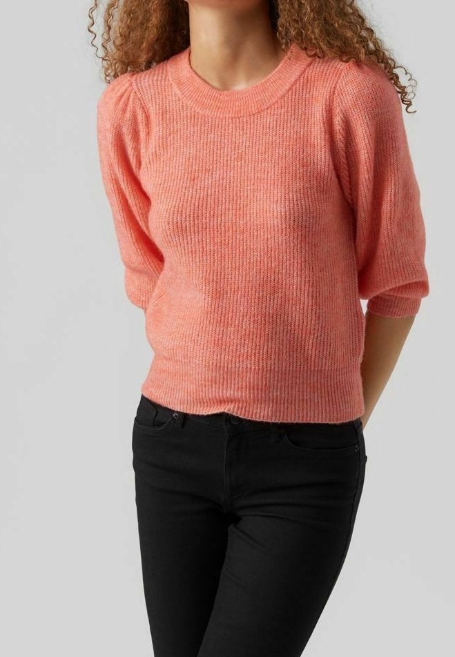 Lichtroze, ribgebreide trui met korte pofmouwen en een ronde hals, in combinatie met zwarte strakke jeans. De textuur lijkt zacht.