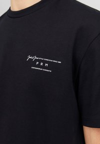 Camiseta de algodón negra con cuello redondo, mangas cortas y un logo impreso en blanco en el lado izquierdo del pecho, mostrando detalles de la marca y el eslogan.