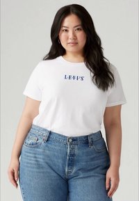 Mulher com cabelo preto comprido, vestindo uma t-shirt branca da Levi's e jeans azuis de cintura alta, de pé contra um fundo claro liso.