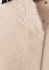 Vaaleanbeige fleece-takki, jossa on ribbineuloskaulus, nepparikiinnitys ja pehmeä pinta. Lähikuva kauluksesta ja napituslistan yksityiskohdista.