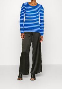 Blå och vit randig långärmad topp i kombination med högmidjade, glänsande svarta vida byxor och svarta öppna klacktofflor.