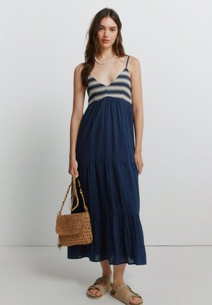 Femme portant une robe midi bleu marine avec un haut en crochet, portant un sac à main tressé et des sandales beiges à bout ouvert sur un fond blanc uni.