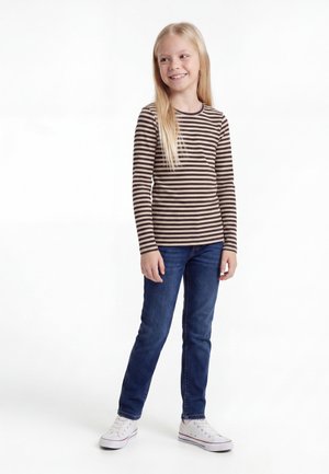 Fille souriante aux longs cheveux blonds, portant un t-shirt à manches longues rayé marron et beige, un jean bleu et des baskets blanches, debout sur fond blanc.
