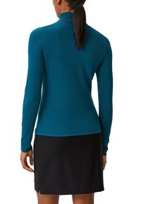 Türkisfarbener Langarm-Top mit hochgeschlossenem Kragen, aus weichem Material, kombiniert mit einem schwarzen Rock mit seitlichen Schlitzen und einer glatten Textur.