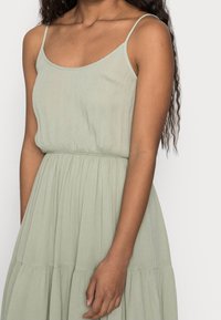 Ljusgrön, ärmlös klänning med smal midja och flytande kjol. Tillverkad av ett strukturerat tyg med subtila veck.