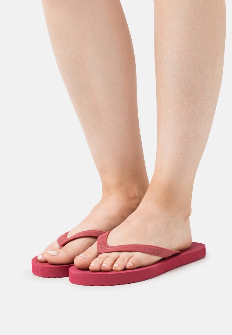 Red rubber flip-flops with a textured V-shaped strap and flat sole, showing the toes and feet of the wearer against a neutral background.