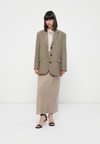 Übergroße olivgrün Blazer mit schwarzen Knöpfen, kombiniert mit einem beigen Midikleid und schwarzen Absatzsandalen. Glatte Stoffqualität mit minimalem Detail.