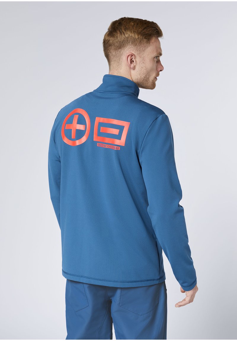 Mann in blauer Jacke und Hose, seitlich stehend, mit großen roten Plus- und Minuszeichen auf dem Rücken der Jacke.