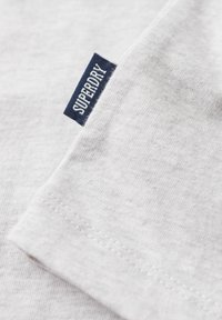 Szara bawełniana koszulka z zaokrąglonym dołem, miękką fakturą i granatową, wplecioną metką z napisem „SUPERDRY” na rękawie.