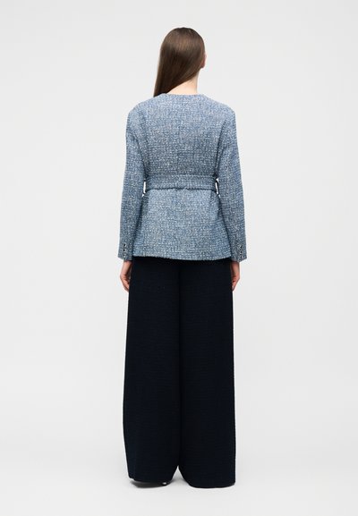 Γυναίκα που στέκεται με την πλάτη, φοράει μπλε καρώ σακάκι με ζώνη και φαρδιά μαύρη παντελόνια, σε απλό ανοιχτό φόντο.