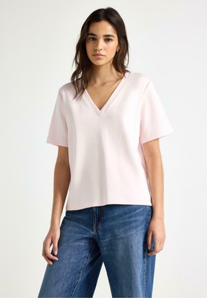 Ung kvinde med mørkt hår iført en lyserød V-hals kortærmet top og blå jeans, stående mod en ensartet hvid baggrund.