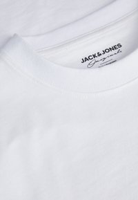 Vit t-shirt med rund halsringning, tillverkad av bomull, som har en mjuk textur och en etikett med "JACK & JONES Originals" tryckt i svart.