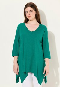 Grüne Tunika mit langen Ärmeln, V-Ausschnitt und asymmetrischem Saum. Hergestellt aus einem weichen, leichten Material mit subtiler Textur und Fall.