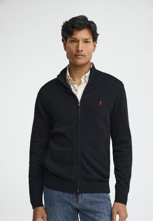 Mann trägt einen schwarzen Reißverschluss-Pullover über einem weißen Hemd und blauen Jeans, blickt nach vorne vor einem einfarbigen hellen Hintergrund.