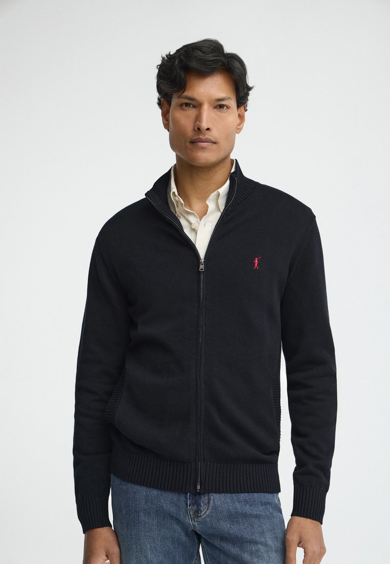 Uomo che indossa un maglione nero con cerniera sopra una camicia bianca e jeans blu, rivolto in avanti contro uno sfondo chiaro e uniforme.