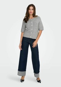 Cardigan gris clair à manches courtes avec boutons, associé à un jean bleu foncé à jambes larges avec des ourlets repliés dans un tissu contrastant. Talons à motifs black and white.