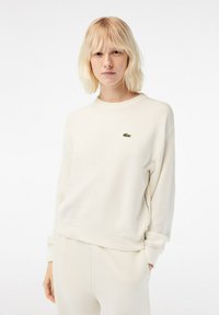 Lichte crèmekleurige sweatshirt met een geribbelde textuur, ronde halslijn en geborduurd groen logo. Heeft een ontspannen pasvorm en mouwtjes met een manchet.
