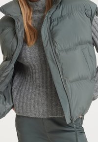 Femme portant un pull long gris côtelé, un gilet matelassé vert foncé avec une fermeture éclair devant, et un pantalon foncé sur un fond uni.