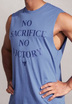 Persona atlética con una camiseta sin mangas azul con el texto "NO SACRIFICIO NO VICTORIA" y un logo de un toro, con las manos apoyadas en las caderas.