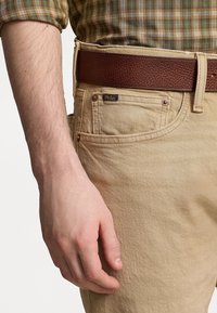 Pantalones de algodón beige con una textura sutil, cinturón de cuero marrón y un logo azul de Polo en el bolsillo trasero. Brazo parcialmente visible.