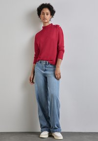 Czerwony sweter w prążki z wysokim kołnierzem i detalami guzika na ramieniu, zestawiony z jasnoniebieskimi spodniami w szerokim kroju i białymi trampkami. Neutralne tło.