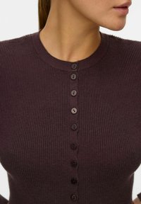 Mörklila ribbad stickad kofta med rund halsringning, prydd med sju matchande knappar fram. Mjuk textur, figurnära design.