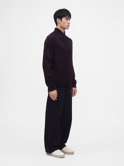 Jeune homme debout de profil portant un pull en maille épais foncé, un pantalon noir large et des baskets blanches sur un fond clair uni.