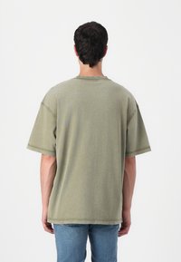 Olive green oversized t-shirt, cotton blend, short sleeves, round neckline, visible stitching details, worn with light blue jeans.