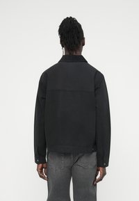 Μαύρο μπουφά ν από τζιν με γιακά, ορατές ραφές και διακοσμητικά κουμπιά στα μανίκια. Η πλάτη διαθέτει μία οριζόντια ραφή.