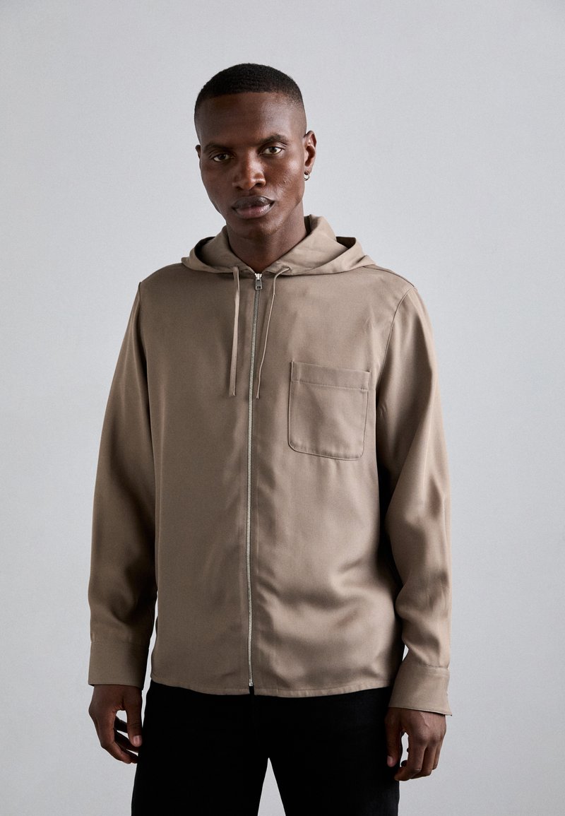 Ανοιχτό καφέ ζακέτα με φερμουάρ και κουκούλα, μπροστινές τσέπες και μακριά μανίκια. Λείο υλικό, μίνιμαλ σχέδιο, φορεμένη πάνω από σκούρα παντελόνια.