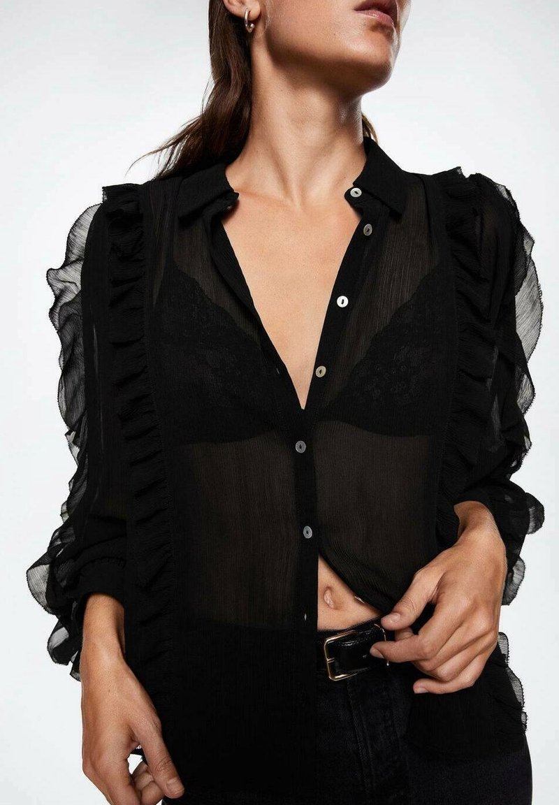 Blusa nera trasparente con maniche a frange, chiusura con bottoni e dettagli in pizzo. Abbinata a jeans scuri, con una cintura nera elegante.