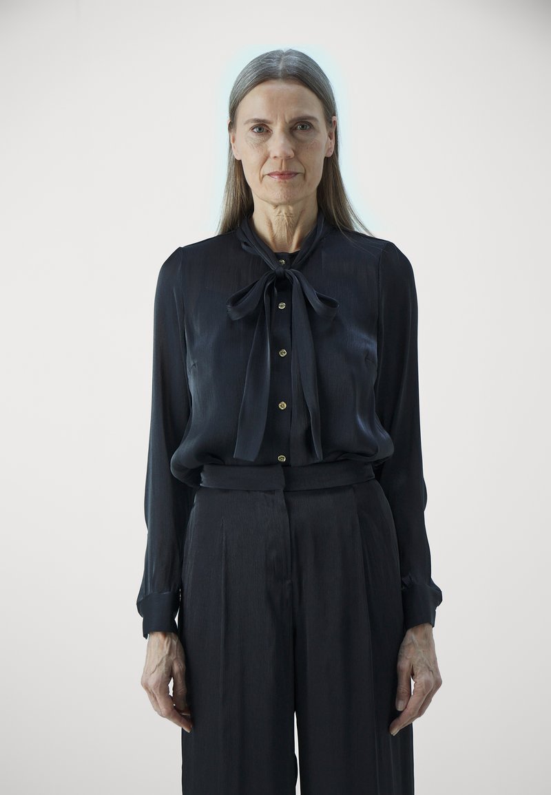 Blusa di seta nera con un fiocco frontale, caratterizzata da un design con bottoni. Abbinata a pantaloni neri a vita alta, entrambi lisci al tatto.