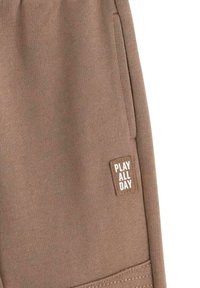 Ruske sweatpants laget av mykt stoff, med en sidelomme og et sydd merke med teksten "LEK HELE DAGEN" i hvitt.