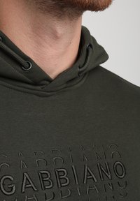 Sudadera verde oliva con un logo de marca texturizado embossado en la tela, que presenta una capucha con cordón y detalles de costura mínimos.