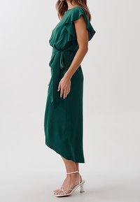 Abito verde al ginocchio con maniche a volant e vita con fiocco. Tessuto morbido con una leggera trama; indossato con sandali argento con cinturini.