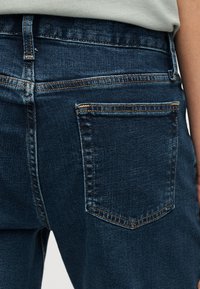 Κοντινό πλάνο πίσω τσέπης και μέσης σκούρου μπλε τζιν με πορτοκαλί ραφές σε άτομο που φοράει ανοιχτό μπεζ τοπ.