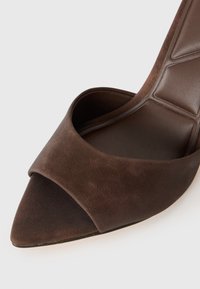 Mule en suede marron foncé à bout pointu avec une large sangle avant et un semelle matelassée sur un fond clair.