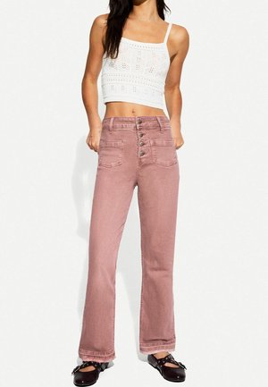 Kobieta nosząca wysokie różowe dżinsy z przodu zapinane na guziki, biały dziergany krótki top i czarne płaskie buty z paskami wokół kostki.