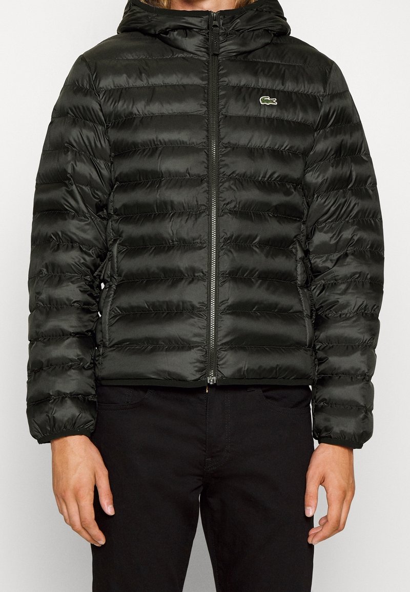 Personne portant une veste noire matelassée à capuche avec fermeture éclair devant et petit logo crocodile vert sur la poitrine, associée à un pantalon noir.