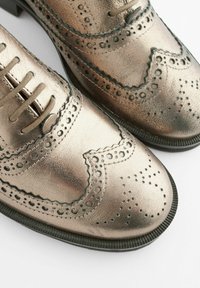 Brogues en cuir bronze métallique avec des perforations décoratives, des bords arrondis et des semelles plates noires. Comprend des lacets et des détails de couture élaborés.