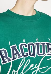 Sudadera de algodón verde con un cuello acanalado, que presenta el texto impreso en blanco y azul marino "RACQUET" y "1983" en una fuente deportiva.