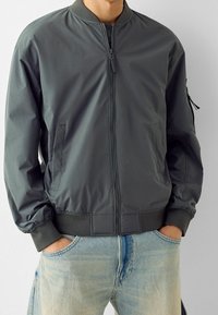 Grå bomberjacka i glatt tyg, med dragkedja, ribbade ärmslut och sidofickor. Bärs över ljusblå jeans.